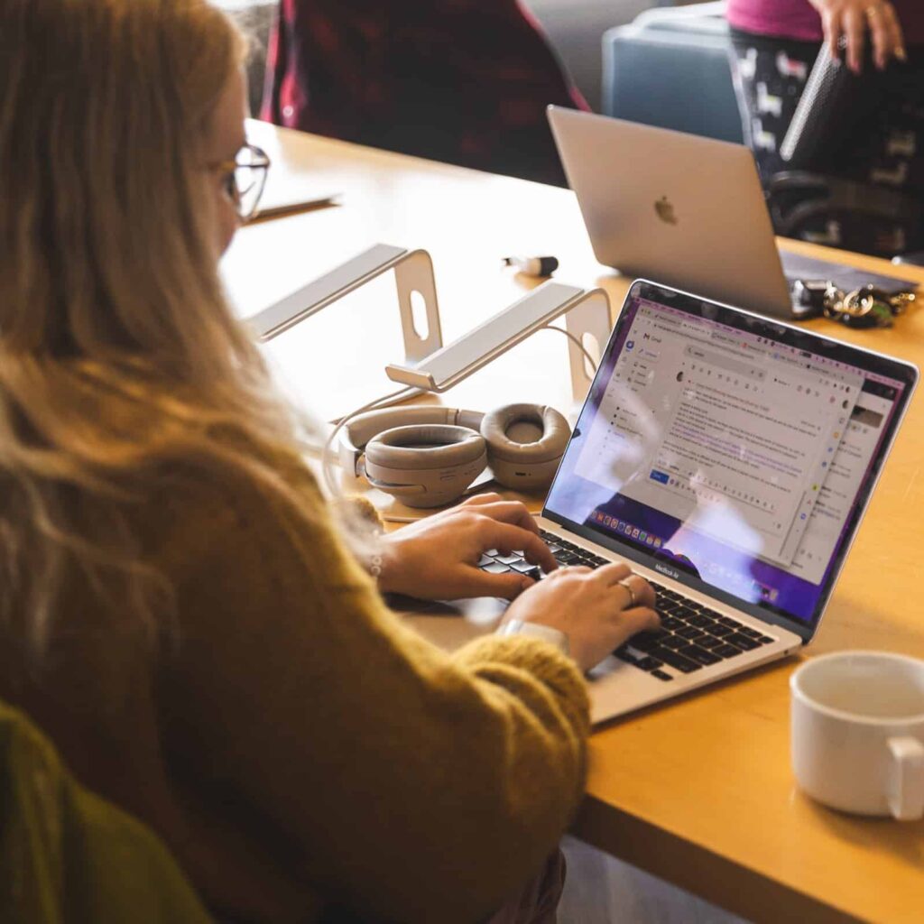 A woman working on a laptop sitting at a desk with headphones and a coffee cup next to her