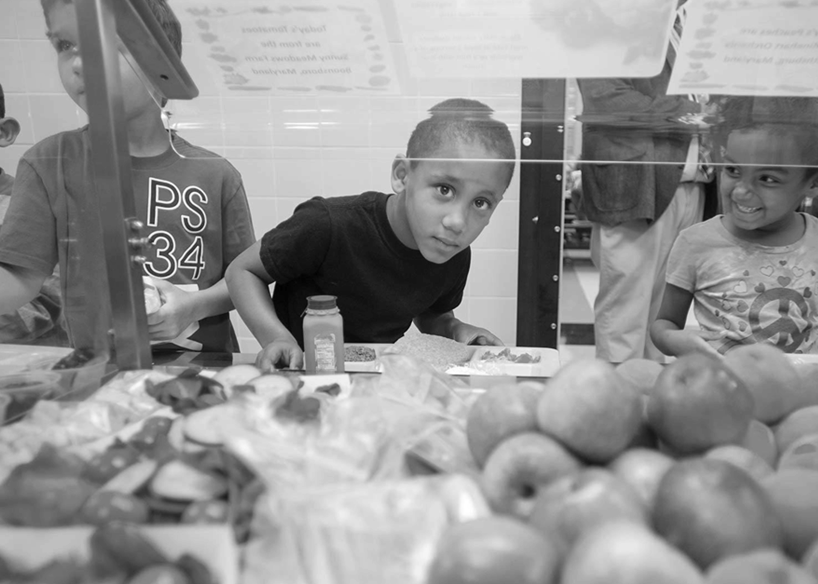 A black and white image of a boy crouching down to look at the camera as he holds a tray of food and waits in line at a cafeteria next to a girl smiling at him