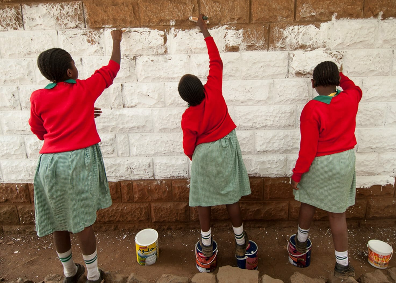 Three girls wearing school uniform face away from the camera and paint a brick wall with white paint