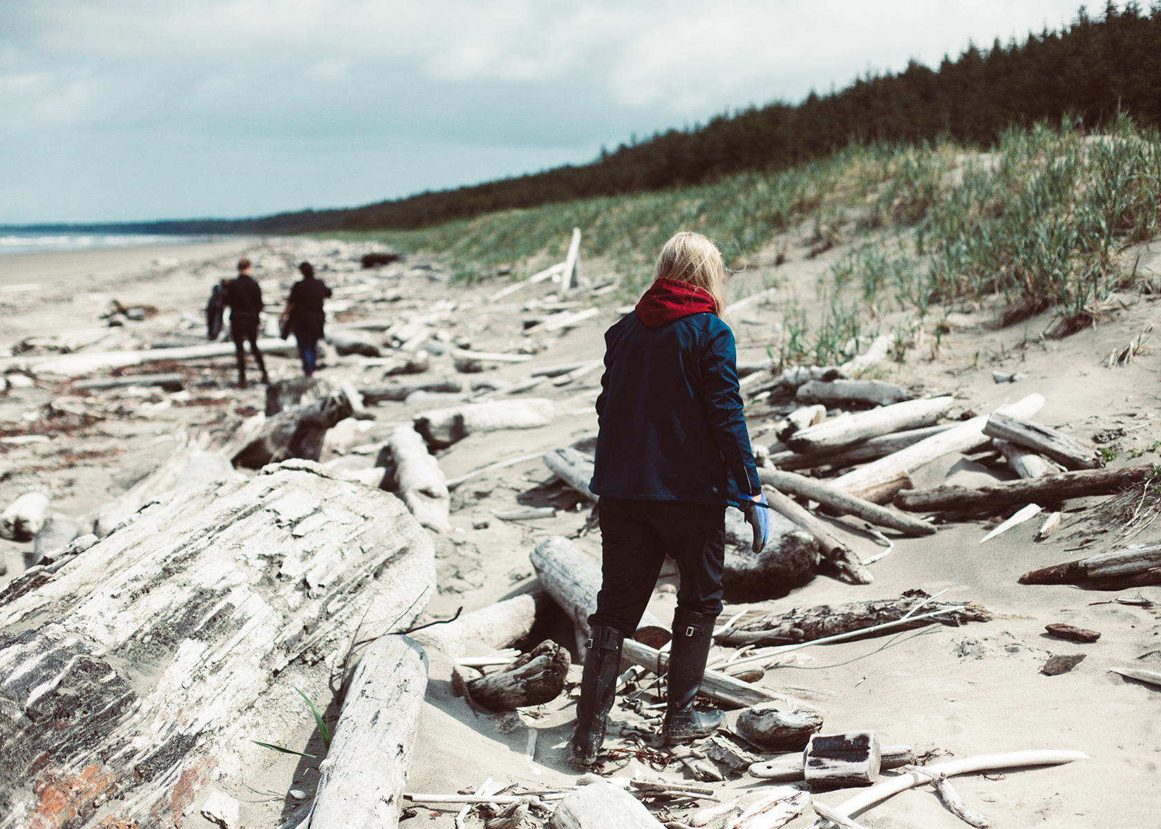 A view from behind of a woman dressed in cold weather clothing and boots walking on a beach covered in driftwood with two firther people walking in the background