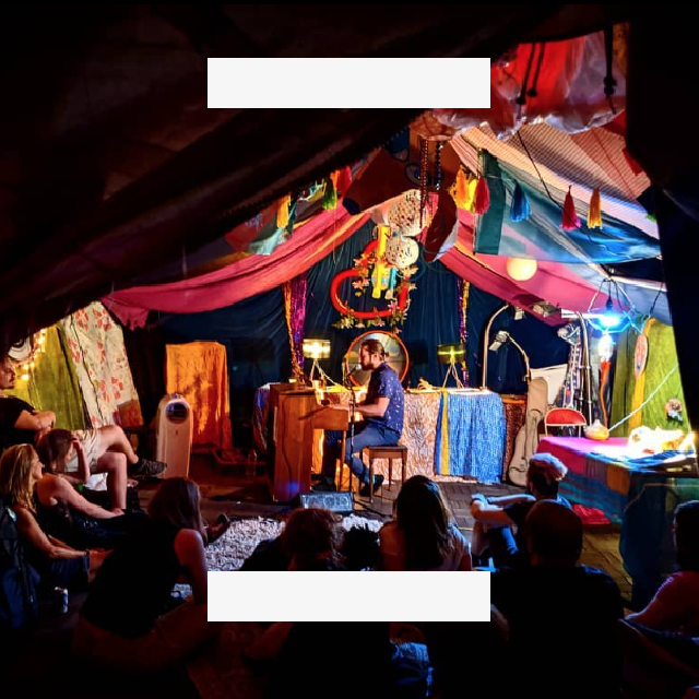 A man plays piano on a stage decorated with colorful fabrics as an audience sits and watches