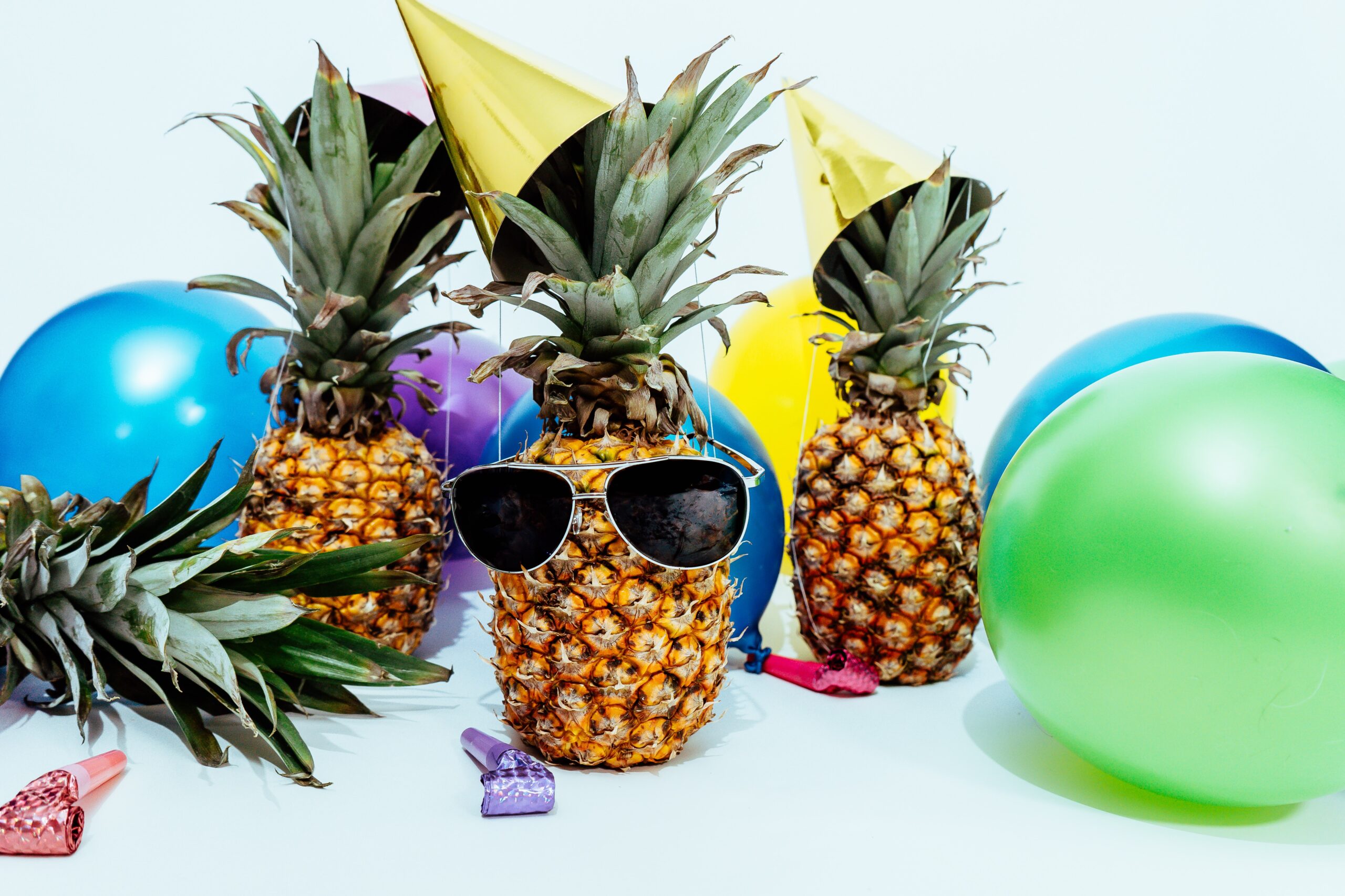 Three pineapples wearing party hats and sunglasses surrounded by balloons