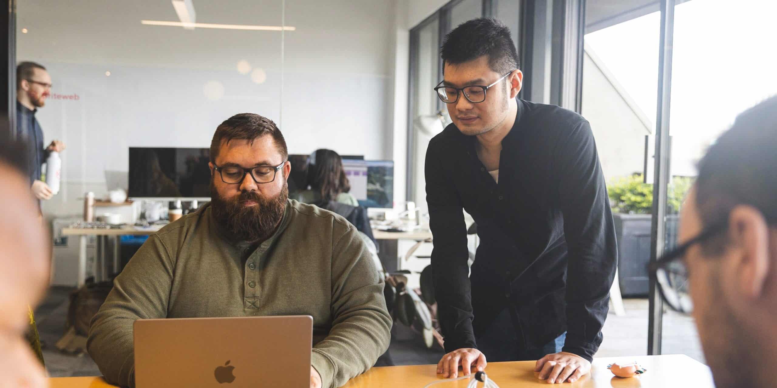 A man sits at a desk and works at a laptop in a busy office while another man stands next to him and looks at his screen