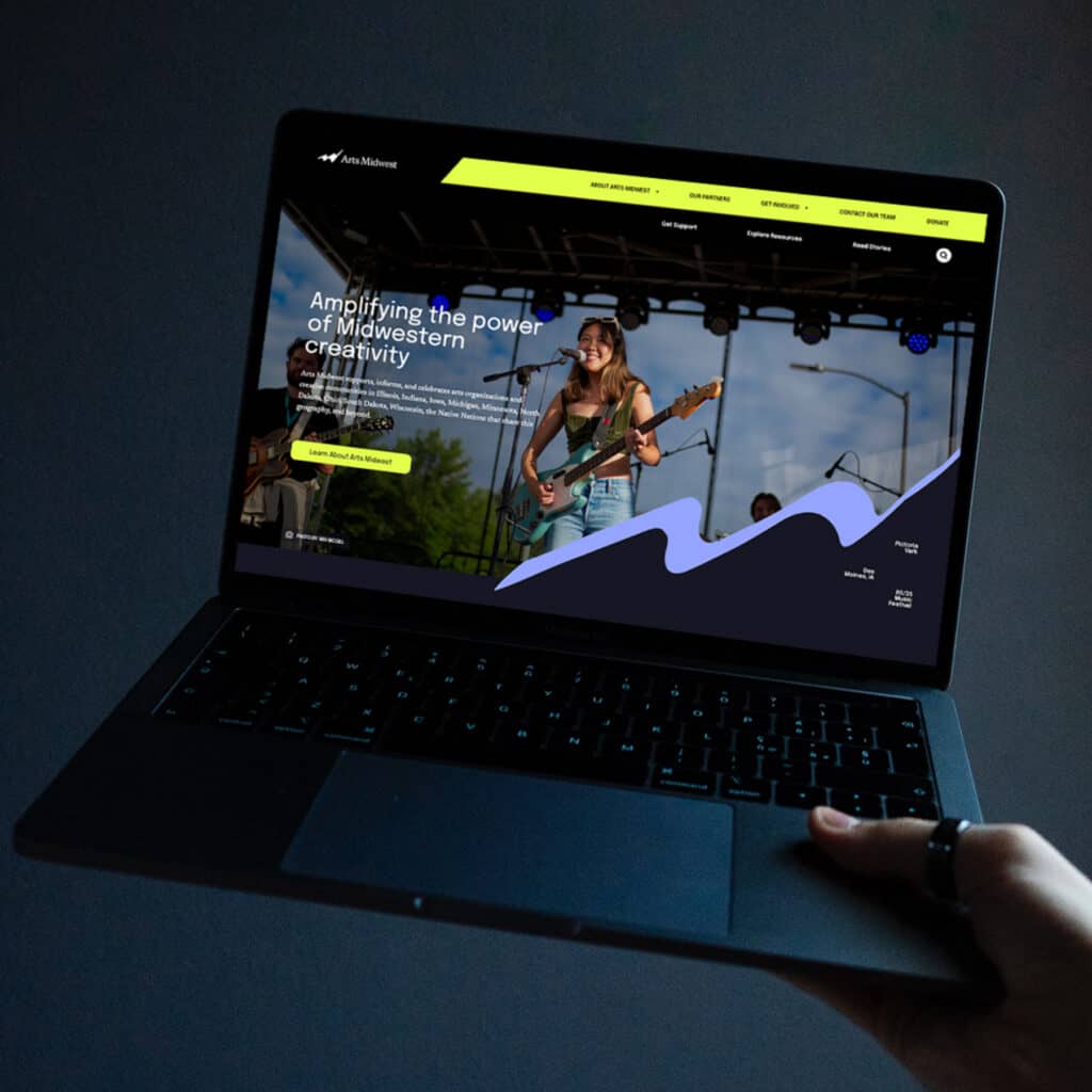 a hand holding a laptop with a full screen of Arts Midwest's homepage