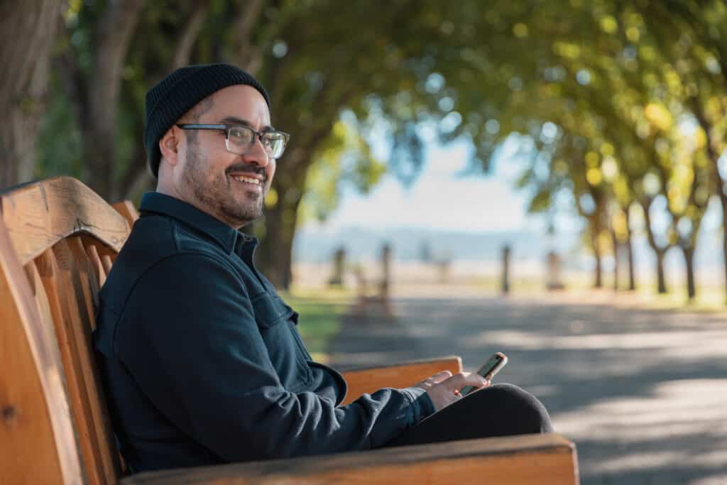 a man in a black shirt and black hat, sitting on a wood bench in a park