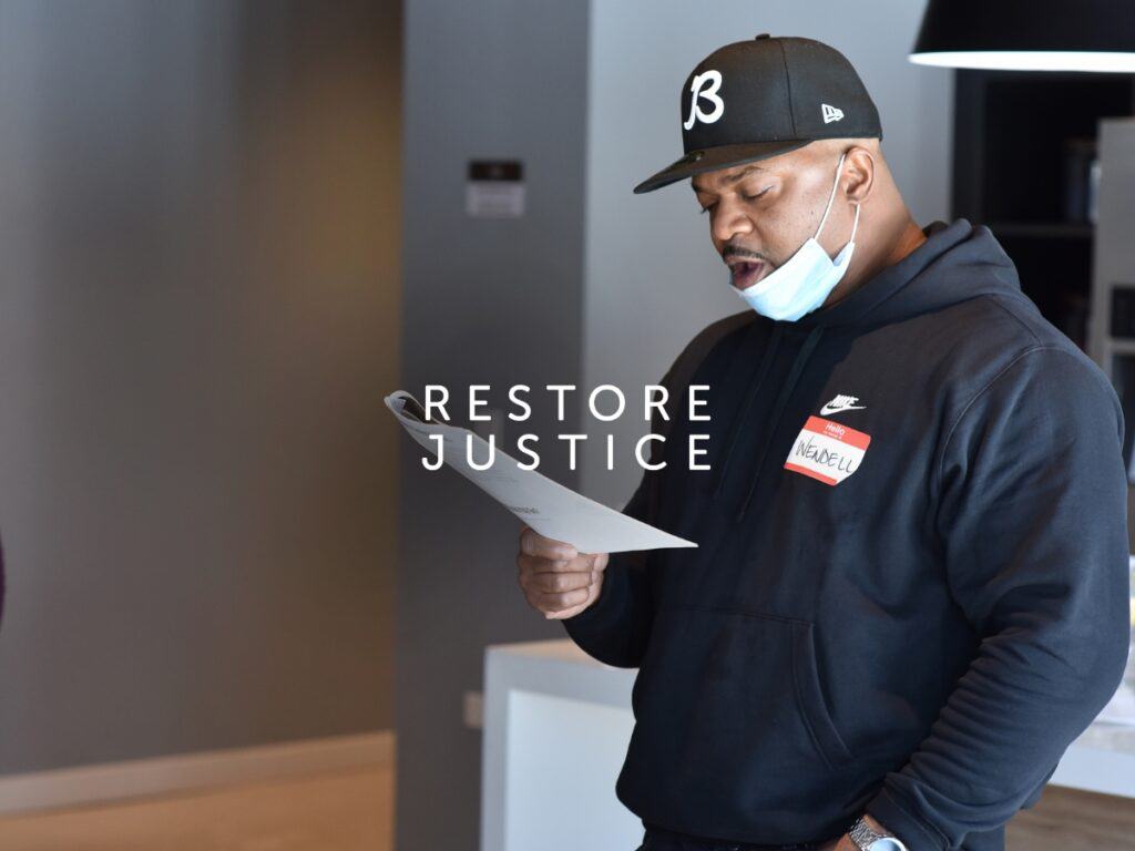 Man in black hoodie and hat reads a document with "RESTORE JUSTICE" text on top of the photo